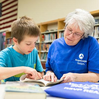 Lady wearing an AmeriCorps Seniors T-shirt reading with a small boy