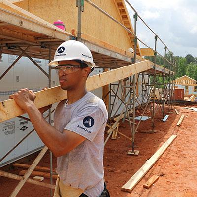 Man wearing an AmeriCorps T-shirt carrying a plank of wood on a building site